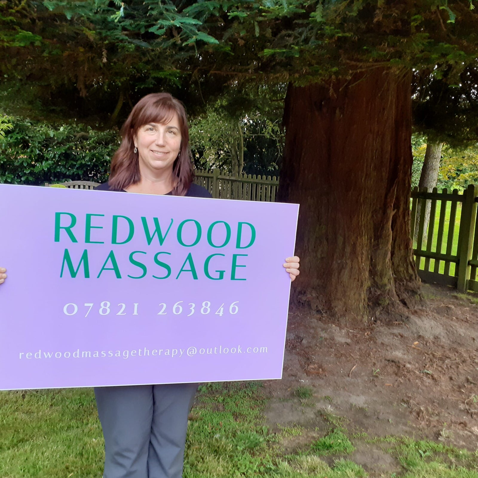 Vicky Noble standing in her garden beside a large redwood trunk, holding a hand-painted lilac board reading Redwood Massage with her phone number and email address.